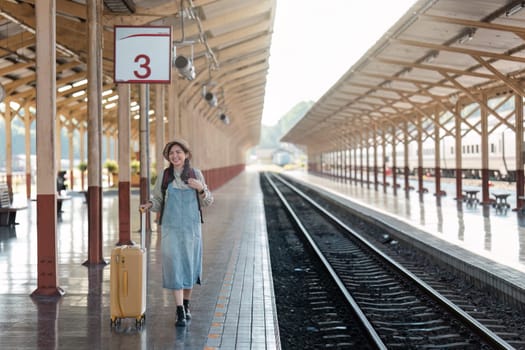 A woman stands with her luggage on a train station platform, ready for her travel adventure. The sunny day highlights the wooden roof structure and railway tracks.