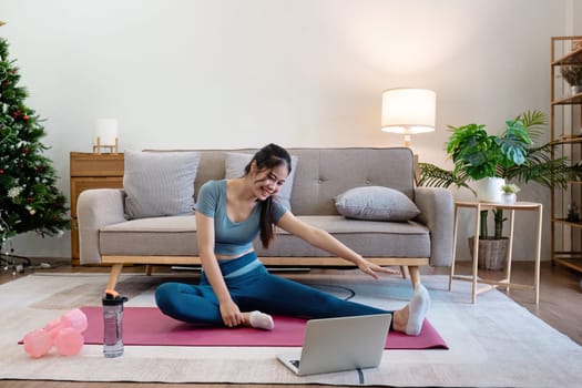 Woman stretching on yoga mat in modern living room with laptop, water bottle, and dumbbells. Cozy and bright interior with sofa, plants, and Christmas tree.