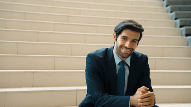 Smiling skilled businessman looking at camera while sitting on stairs. Young professional project manager smile at camera while holding mobile phone at outdoor with blurred background. Exultant.