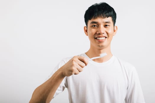 Smiling Asian young man emphasizes dental health while brushing teeth. Studio shot isolated on white background, promoting oral care and dentistry.