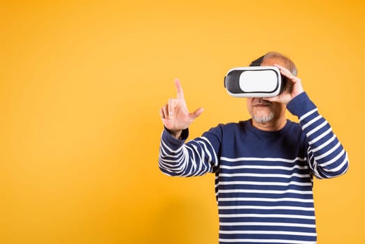 Portrait Asian smiling old man pointing at something in a virtual reality glasses headset studio shot isolated yellow background, Excited happy senior man pensioner using VR play simulation game