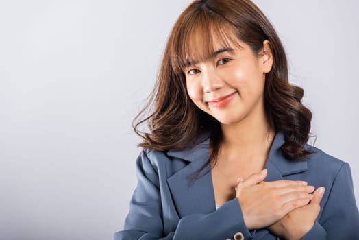 A happy business woman in casual attire holds her hands close to her chest, meditating and smiling. Studio shot isolated on a white background, portraying the concepts of belief, faith, and gratitude