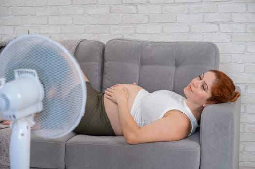 Pregnant woman suffers from heat and lies on the sofa near the electric fan