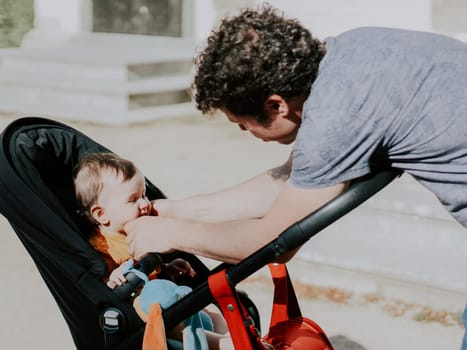 Caucasian young and handsome man dad touches his hands to the cheeks of a little daughter sitting in a stroller with her eyes closed from the bright sun in a public park, close-up side view. Concept of fatherhood, dads, family vacation, spring walk, dad care.
