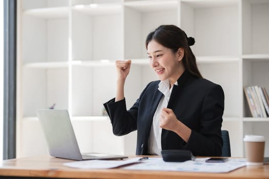 Professional businesswoman working on a laptop in a modern office, celebrating success with a fist pump, showcasing productivity and achievement.