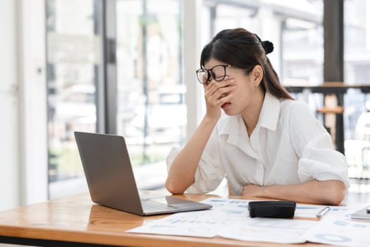 Businesswoman feeling stressed while working on a laptop at her desk, surrounded by documents in a modern office setting.