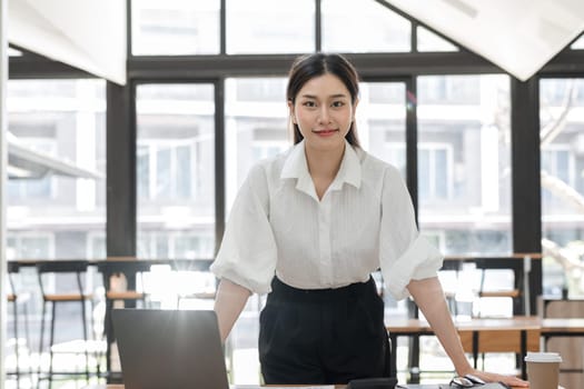 Professional business woman working in a modern office environment, standing confidently with a laptop and documents.