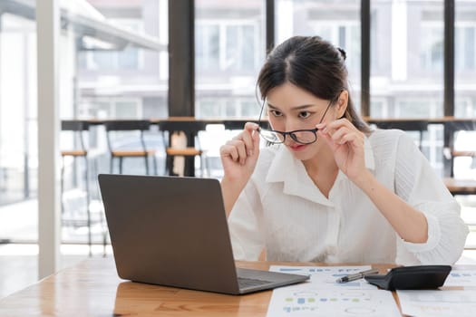 Businesswoman feeling stressed while working on a laptop in a modern office, analyzing financial reports, and struggling with workload.