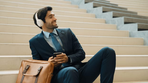 Professional smart business man listening music while lie on stairs. Smart manager happy while receive good news or getting project from headphone. Investor wear suit and bag while sitting. Exultant.