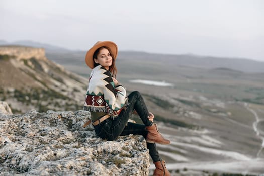 Serene woman in cozy outfit sitting on mountain summit with boots and hat enjoying the view