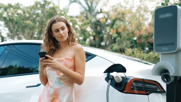 Young woman use smartphone to pay for electricity at public EV car charging station in nature. Modern environmental and sustainable automobile transportation lifestyle with EV vehicle. Synchronos