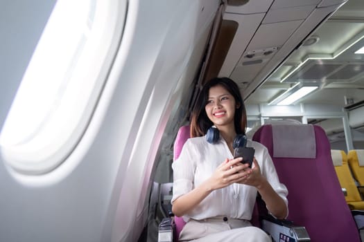 Professional businesswoman traveling by airplane, smiling and using smartphone with headphones around neck, representing modern corporate travel and in-flight comfort.