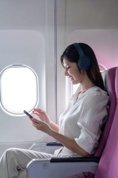 Businesswoman traveling by airplane, using smartphone and wearing headphones, seated by window, enjoying modern air travel.