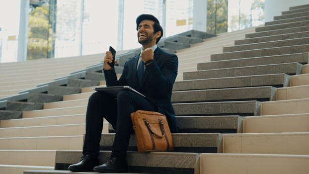 Successful business man celebrate successful project while sitting at stairs. Smart project manager getting new gob, getting promotion, increasing sales while calling friends by using phone. Exultant.