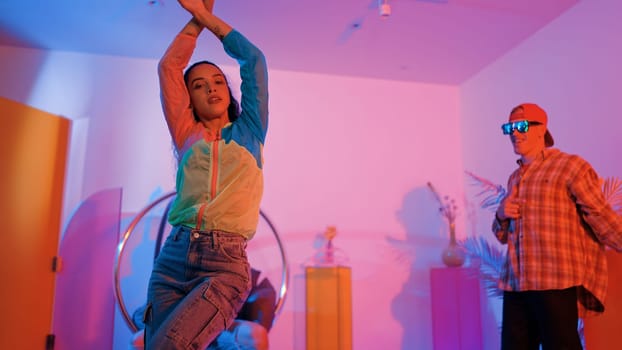 Attractive happy dancer looking at camera while smart woman moving at rhyme with neon light. Professional hispanic performer break dancing while wearing colorful cloth with diverse friend. Regalement.