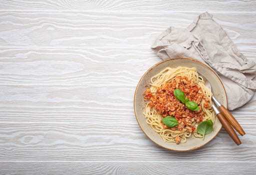 Plate of spaghetti bolognese, topped with fresh basil leaves, sits on a white wooden table with a linen napkin