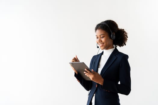 A professional female call center employee wearing a headset, smiling, and using a tablet in a modern office environment.