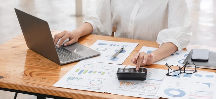 Focused business woman analyzing financial data with a laptop and calculator, surrounded by charts and documents in a modern office environment.