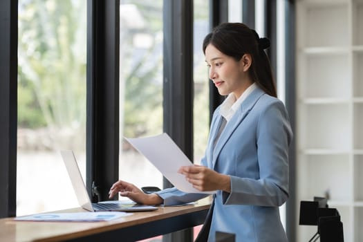 Business woman in formal attire working on a laptop, analyzing documents in a modern office with large windows.