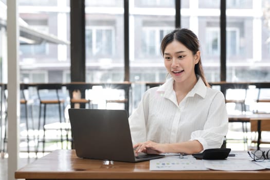 Business woman working on a laptop in a modern office, smiling and engaged in productive work with documents and a calculator on the desk.