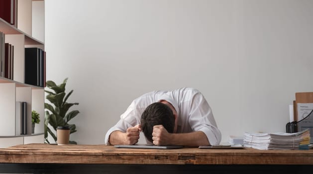 A young businessman sits at his desk with his head down, expressing disappointment and frustration at work. The modern office setting highlights stress and burnout.