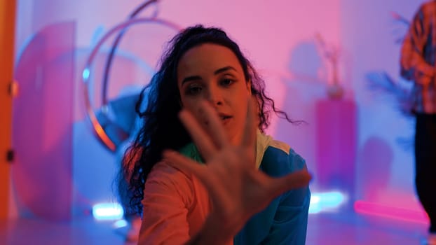 Attractive happy dancer looking at camera while smart woman moving at rhyme with neon light. Professional hispanic performer break dancing while wearing colorful cloth with diverse friend. Regalement.