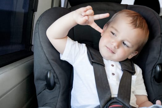 Little boy sitting in a car seat in the car making the victory hand. Peace sign.