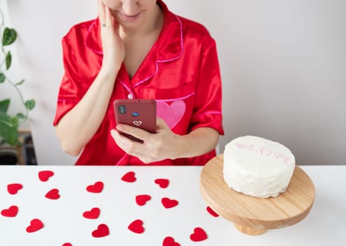A girl in a red shirt uses a smartphone while a cake sits on a wooden stand, surrounded by heart-shaped confetti on the table