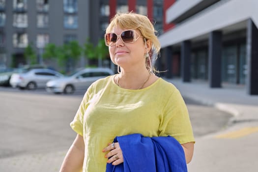 Portrait happy elegant mature woman 50 years old in sunglasses outdoor, urban background. Smiling beautiful blonde female in yellow looking at camera in city. Age, lifestyle, beauty, fashion concept