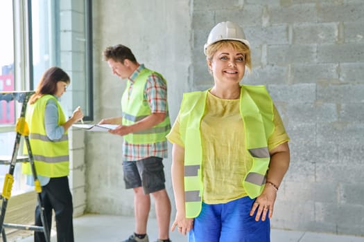 Female designer managing finishing decoration in safety helmet vest looking at camera with team workers in background in cement room of office. Industrial workers construction repair work concept