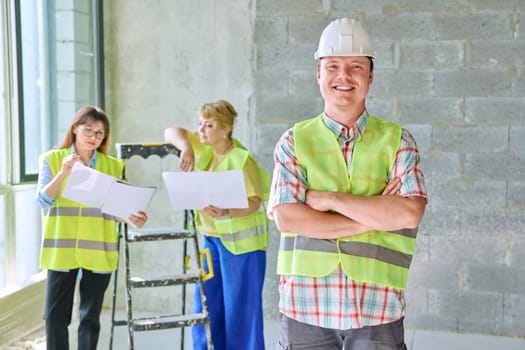 Young male industrial worker in safety helmet vest at construction site inside, finishing work on construction site. Repair, renovation, electrical, engineering, plumbing works, decorating, teamwork