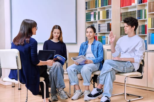 Group of teenage students sitting with female mentor teacher psychologist counselor social worker inside classroom library office. High school education teamwork learning behavior adolescence concept