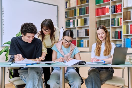 Group of teenage students sitting at desk with female mentor teacher study together prepare for tests inside classroom library office. High school, education, teamwork, learning, adolescence concept