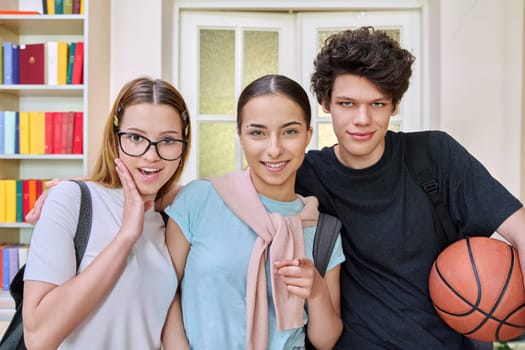 Three cheerful smiling teenagers friends students looking at camera inside high school classroom campus. Adolescence, friendship, education, 17-18 years old students, teenage concept