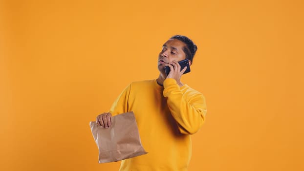 Indian man holding thermal backpack answering phone, fulfilling clients orders, isolated over studio background. Food delivery service employee talking with customer during telephone call, camera B