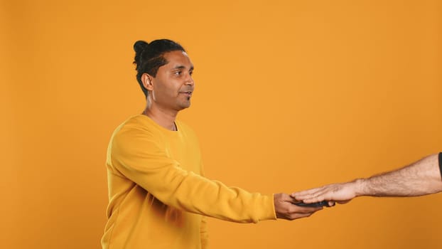 Indian pizza delivery worker getting takeaway order out of thermal backpack, giving it to client paying with card. Courier offering lunch paper bag and POS to customer, studio background, camera A