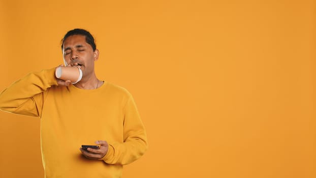 Indian man enjoying fresh coffee from disposable paper cup early in the morning to wake up and be energized. Person drinking hot beverage from recycled takeaway cup, studio background, camera A