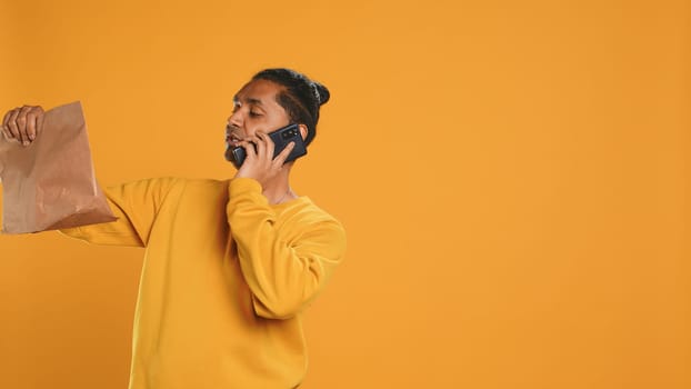 Indian man holding thermal backpack answering phone, fulfilling clients orders, isolated over studio background. Food delivery service employee talking with customer during telephone call, camera A