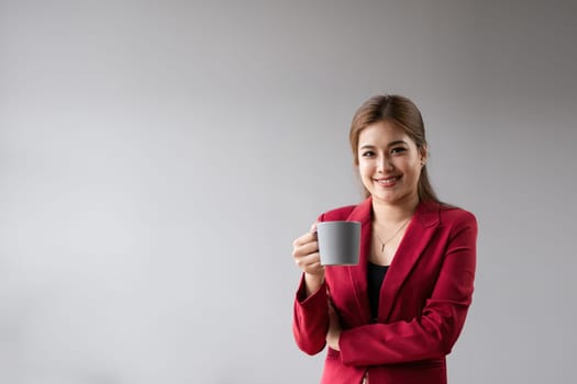 Confident business woman in red blazer holding coffee mug, smiling, and standing against minimalist background.