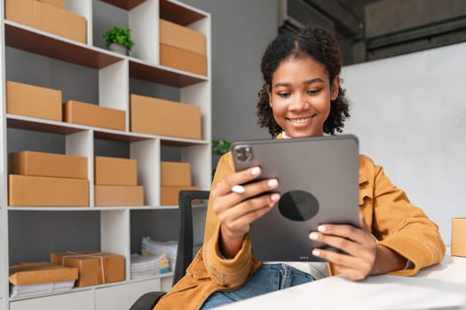 Young female entrepreneur using a tablet to manage her online business in a modern office setting with shipping boxes.