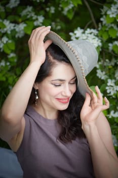 Elegant Woman Posing With Hat in Blossoming Garden. Smiling woman in a dress playfully tipping her hat among spring blossoms. The concept of fashion, self care