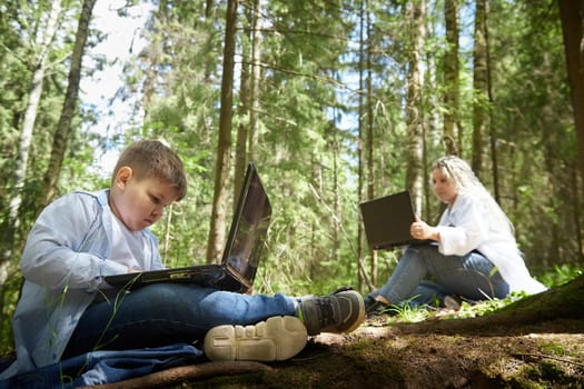 Mother and son with a laptops in the forest in summer. Fat young smart teenage boy and woman working with modern IT technologies in nature