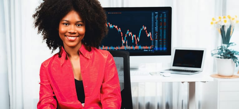 Analytical young African American businesswoman, a specialist in successful stock exchange trading, against dynamic data graph displaying marketing trend analysis on screen. Tastemaker.