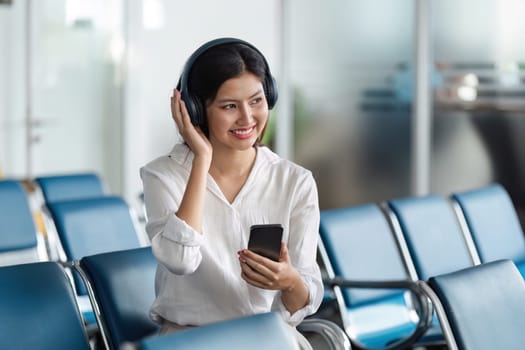 A young woman in an airport terminal, smiling and listening to music with headphones while holding a smartphone, representing the travel airplane concept.