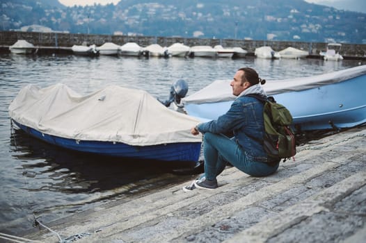 A man with a green backpack sitting on steps by the lake. Covered boats float in the background on a misty day, creating a serene, contemplative atmosphere.