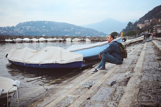 A man sits by the lakeside with docked boats, gazing at the surrounding mountains and reflecting on the peaceful atmosphere. Perfect for themes of travel, relaxation, and contemplation.