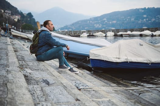 Man sitting on a dock by a marina, looking contemplative. Mountains in the background with moored boats, calm water, and a peaceful atmosphere.