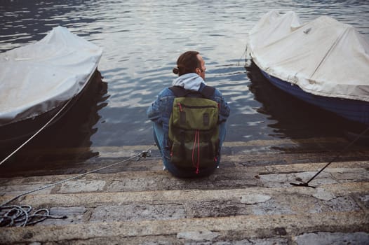 Rear view of a man with a backpack sitting on stone steps near water, flanked by two boats. The scene exudes tranquility and contemplation.