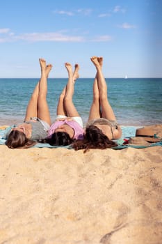 Three young women lying on a tropical beach, stretching their slender legs. Blue sea in the background. Summer vacation concept.