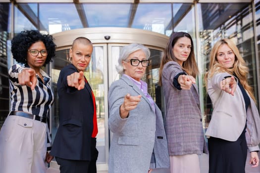 Five multiracial businesswomen standing, side by side, looking at the camera smiling and pointing their fingers. Suitable for team concepts, friendship and diversity.